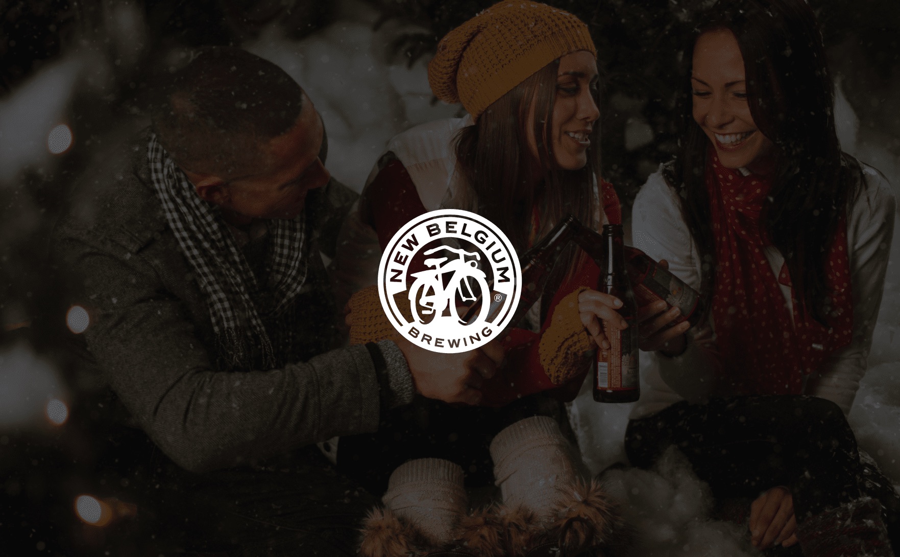 White New Belgium Brewing logo with man and two women toasting beer bottles in a winter scene in the background