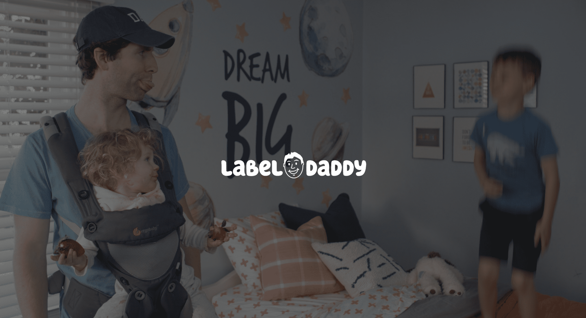 Label Daddy Featured Image White Label Daddy logo with background of Daddy doing a raspberry at boy standing on bed while carrying a girl in a chest carrier who is holding strawberries