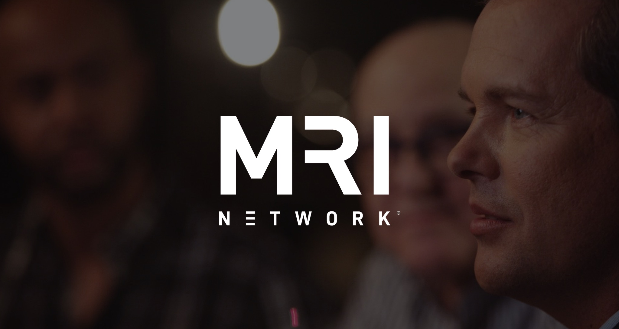 White MRI Network logo with dimmed background closeup of man talking amongst others