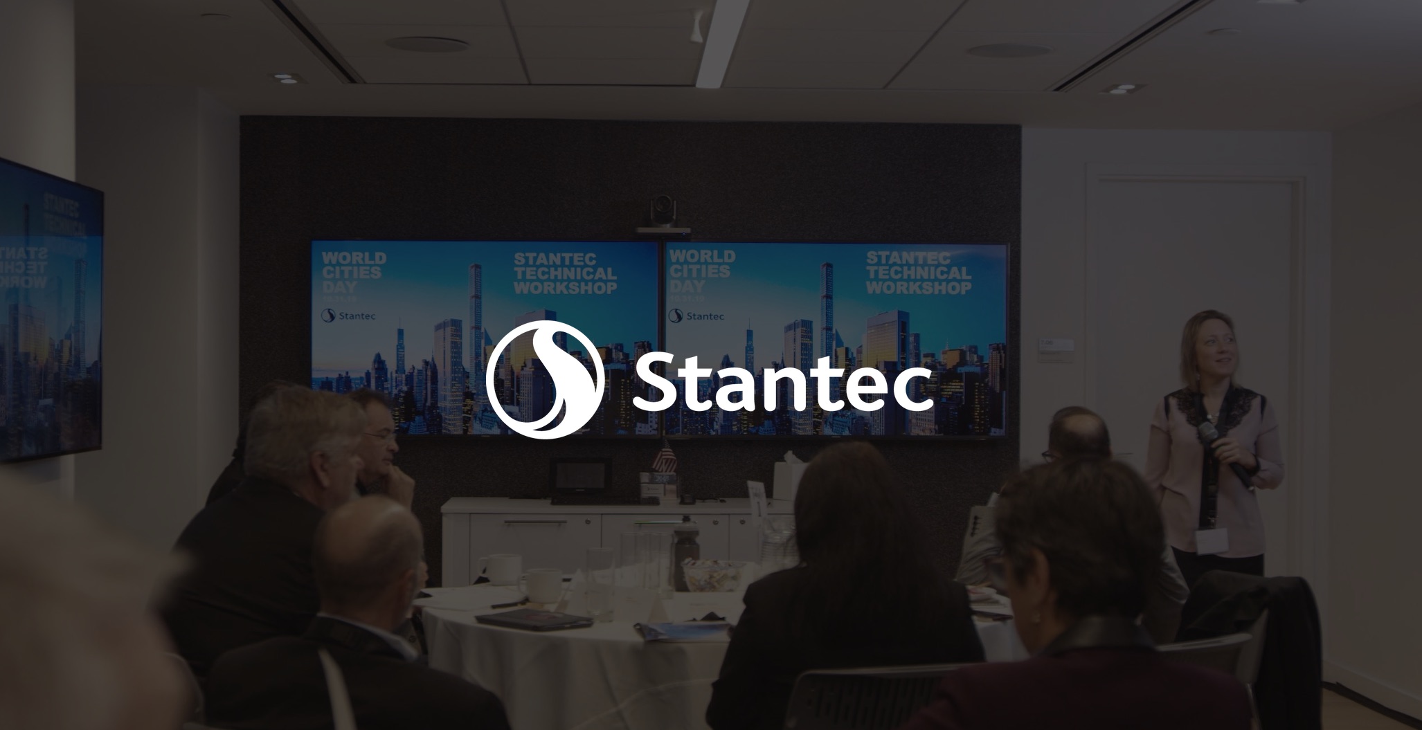 White Stantec logo over dimmed background of Live Video Streaming Event with many people in a room with video conferencing displays