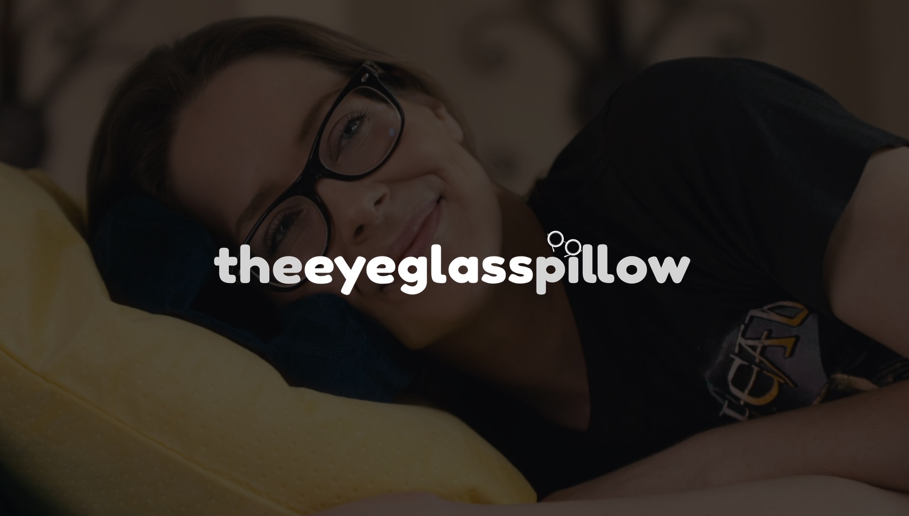 White The Eyeglass Pillow logo by LaySee Pillow with dimmed background of woman wearing black t shirt laying on her side smiling