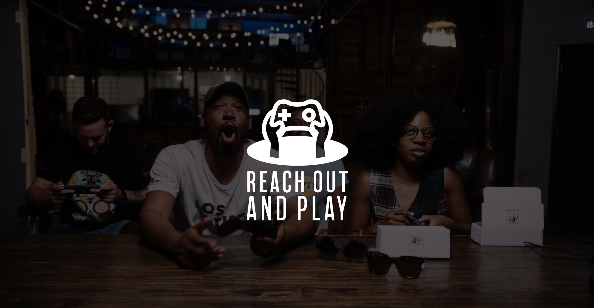 White Reach Out and Play icon with dimmed background of three men and a woman playing video game with video game controllers with two shades and white boxes on the table in front of them