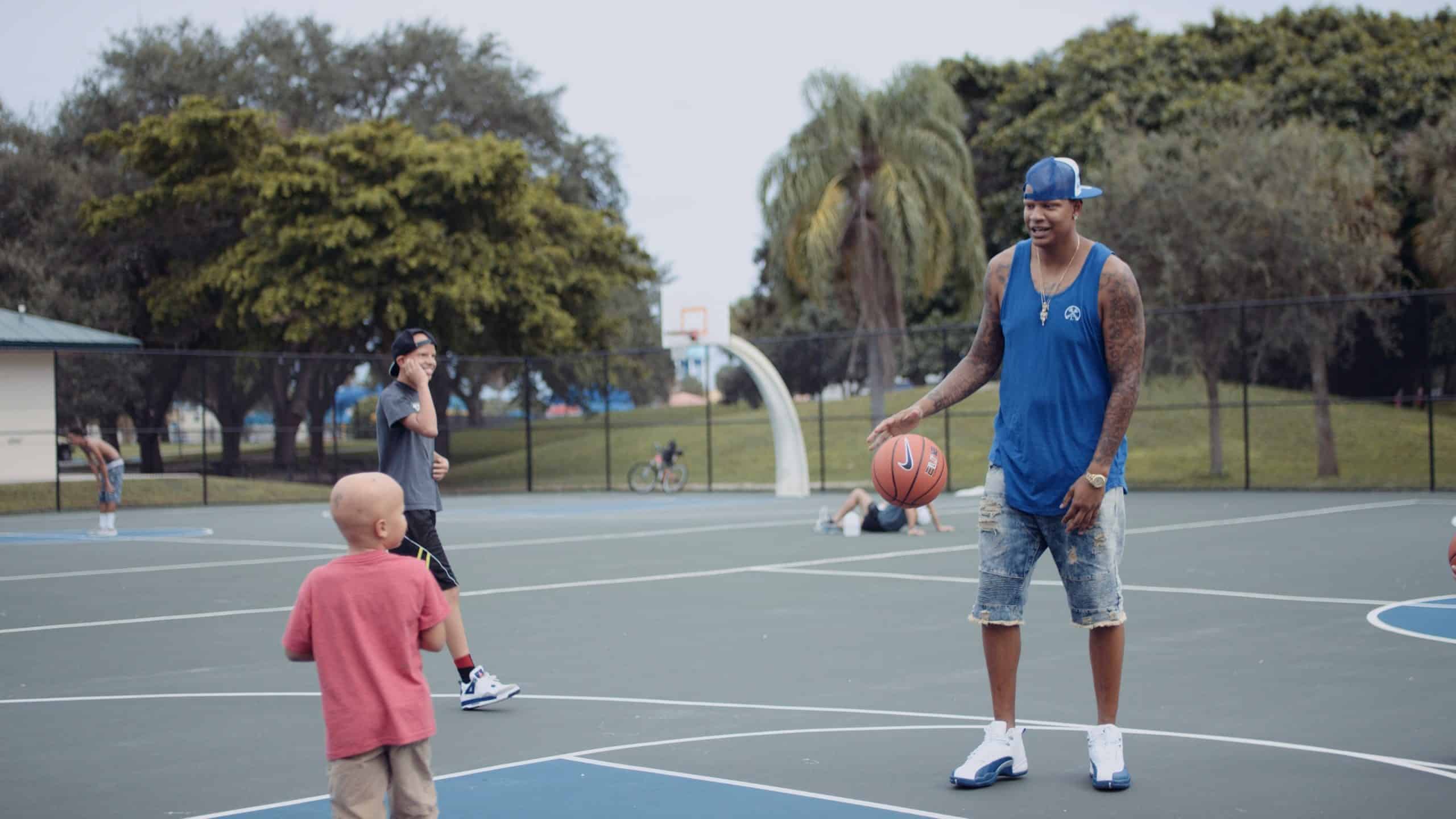 Charlie V What is Alopecia? Still of Charlie V sitting wearing a blue top and baseball cap on backwards playing on a basketball court with a boy and a teenager