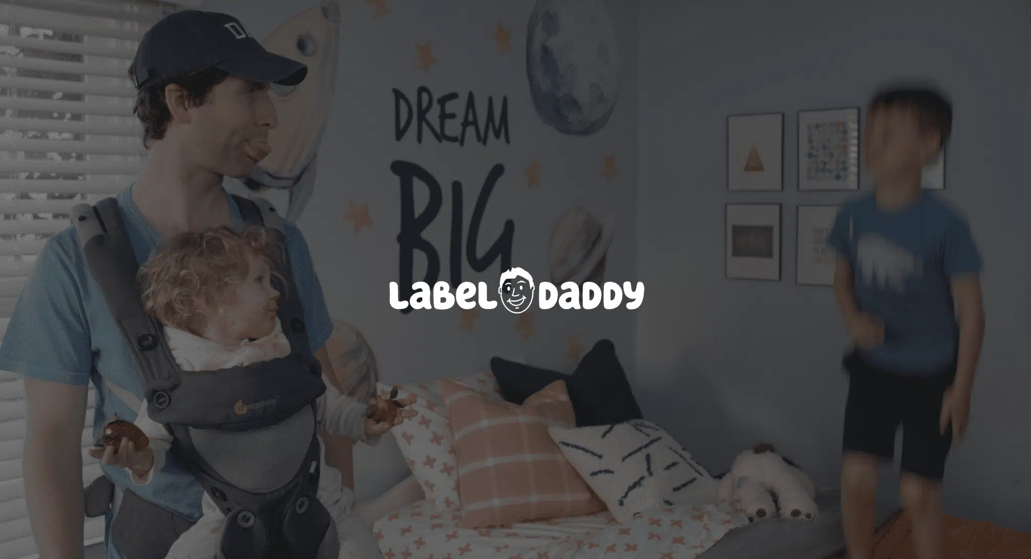 IU C&I Studios Page Label Daddy Featured Image White Label Daddy logo with background of Daddy doing a raspberry at boy standing on bed while carrying a girl in a chest carrier who is holding strawberries