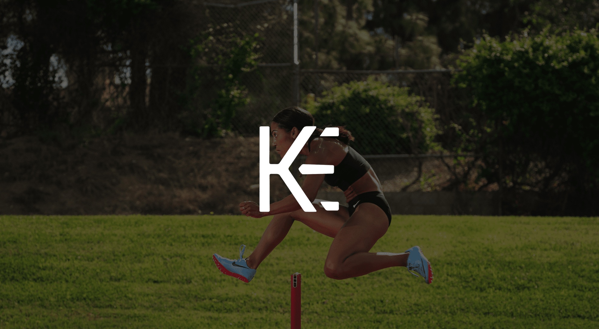 Keyword: Professional photography services .White KE logo against a dimmed background side view of a female track athlete jumping a hurdle