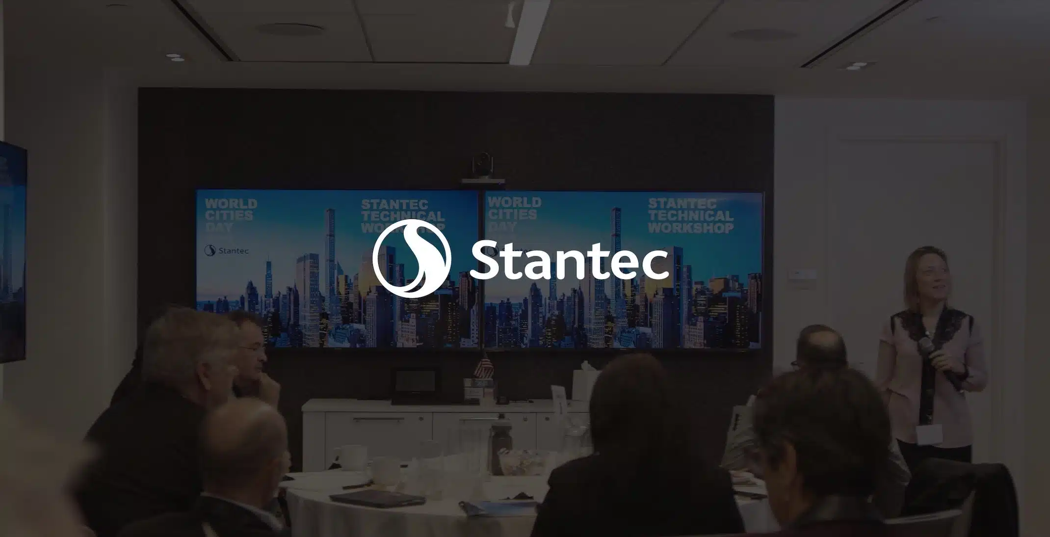 White Stantec logo over dimmed background of Live Video Streaming Event with many people in a room with video conferencing displays. Live streaming