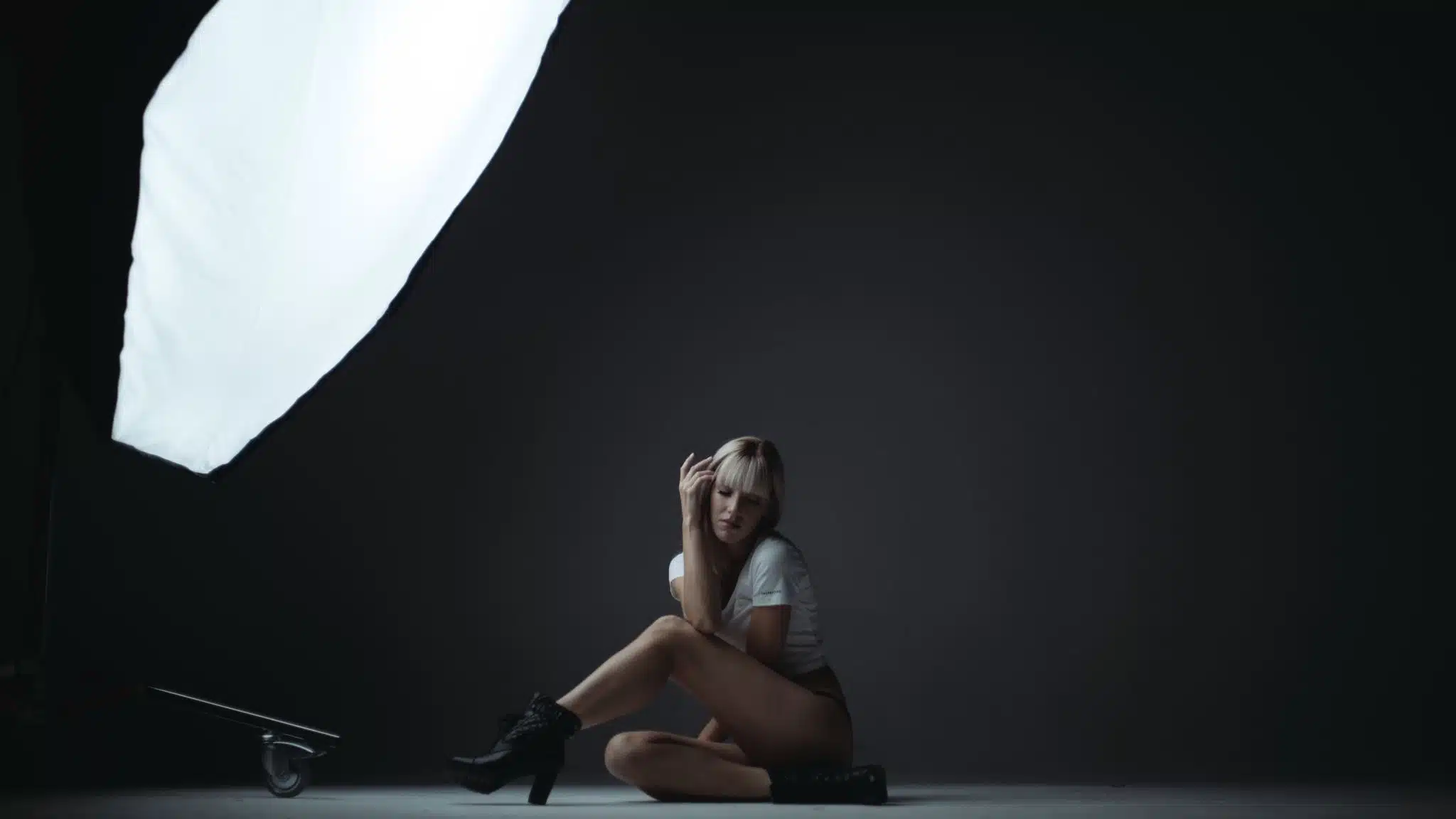 Woman with blond hair sitting on the ground posing for the camera looking down next to lighting equipment