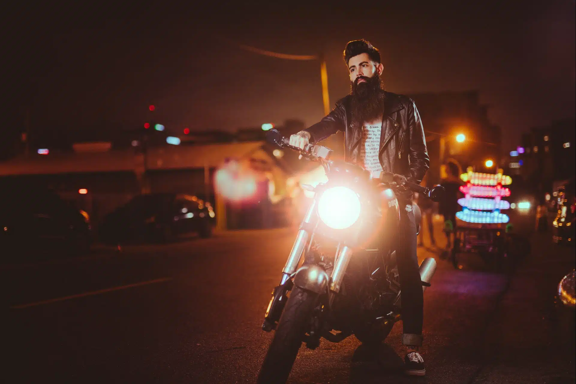 Man with a long beard wearing a leather jacket on a motorcycle at night with headlight on waiting on the street