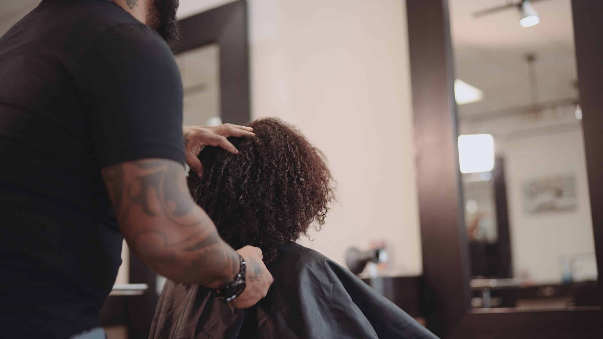 All-Dolled-Up 8K All Dolled Up Month 1 View from behind of woman with brown fizzy hair with a male hairdresser standing behind her working on her hair