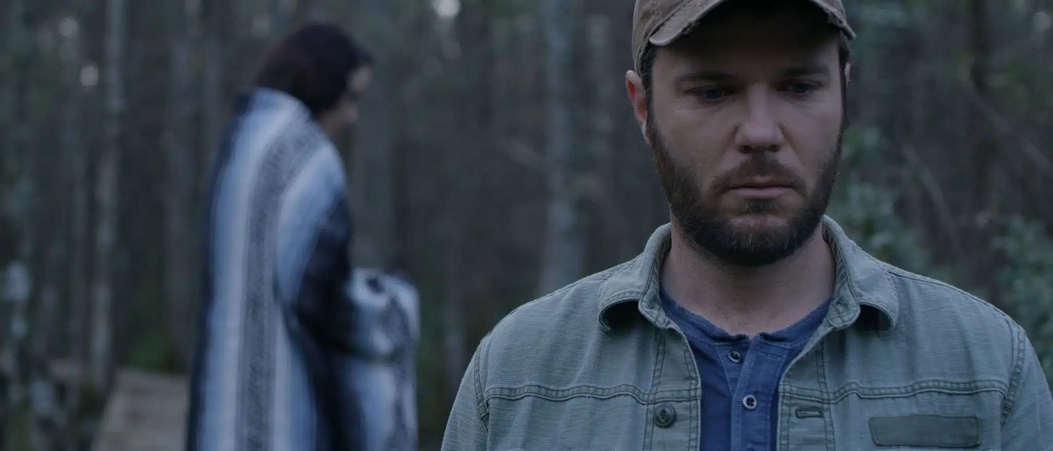 Man wearing a brown ball cap in the forefront looking down while a woman stands in the background with a blanket around her shoulders.