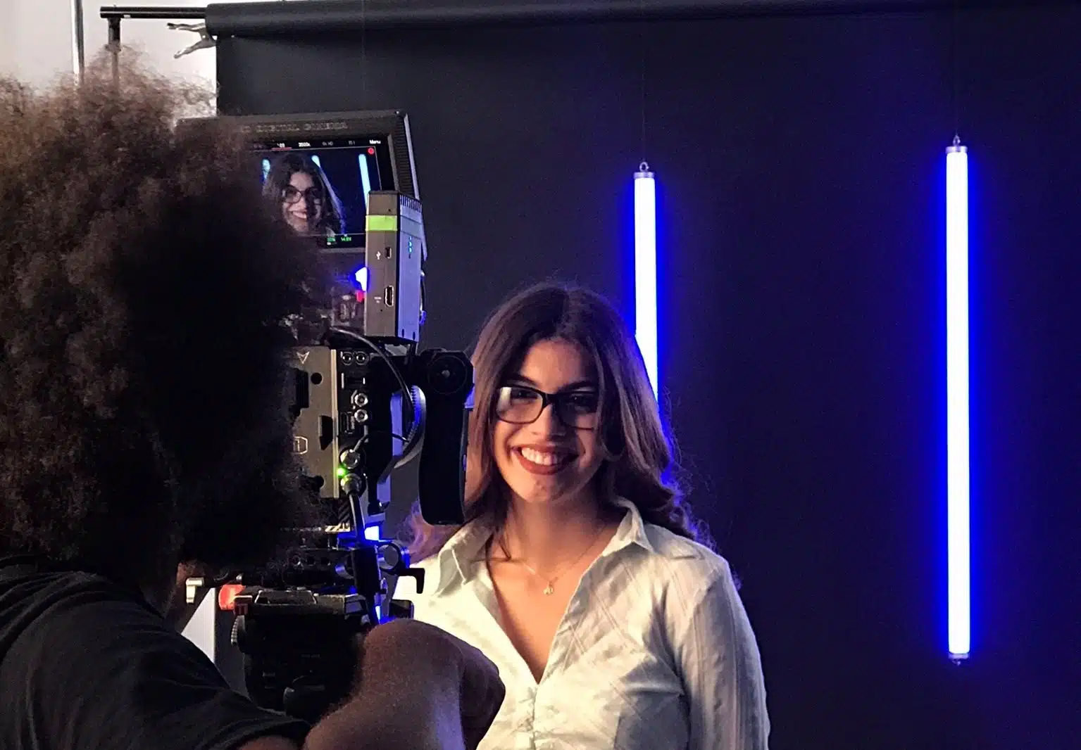 View from behind of male crew member filming a woman with long brown hair wearing a white top smiling