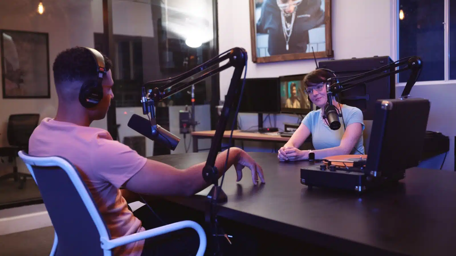 Side profile of a man wearing black headphones and a peach colored t shirt with a woman by microphones