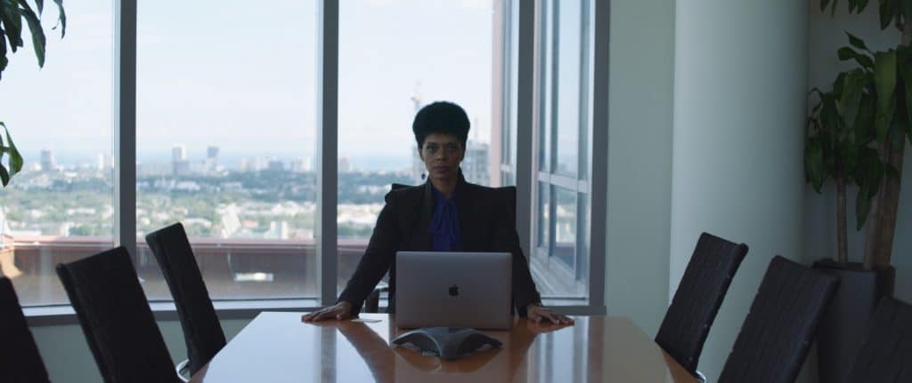 Woman sitting at an empty table in a conference room behind a laptop posing for the camera with hands on the table