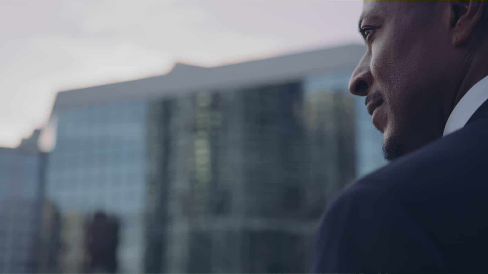 View from behind of an African American man wearing a blue suit with white shirt looking off to the side with a building in the background
