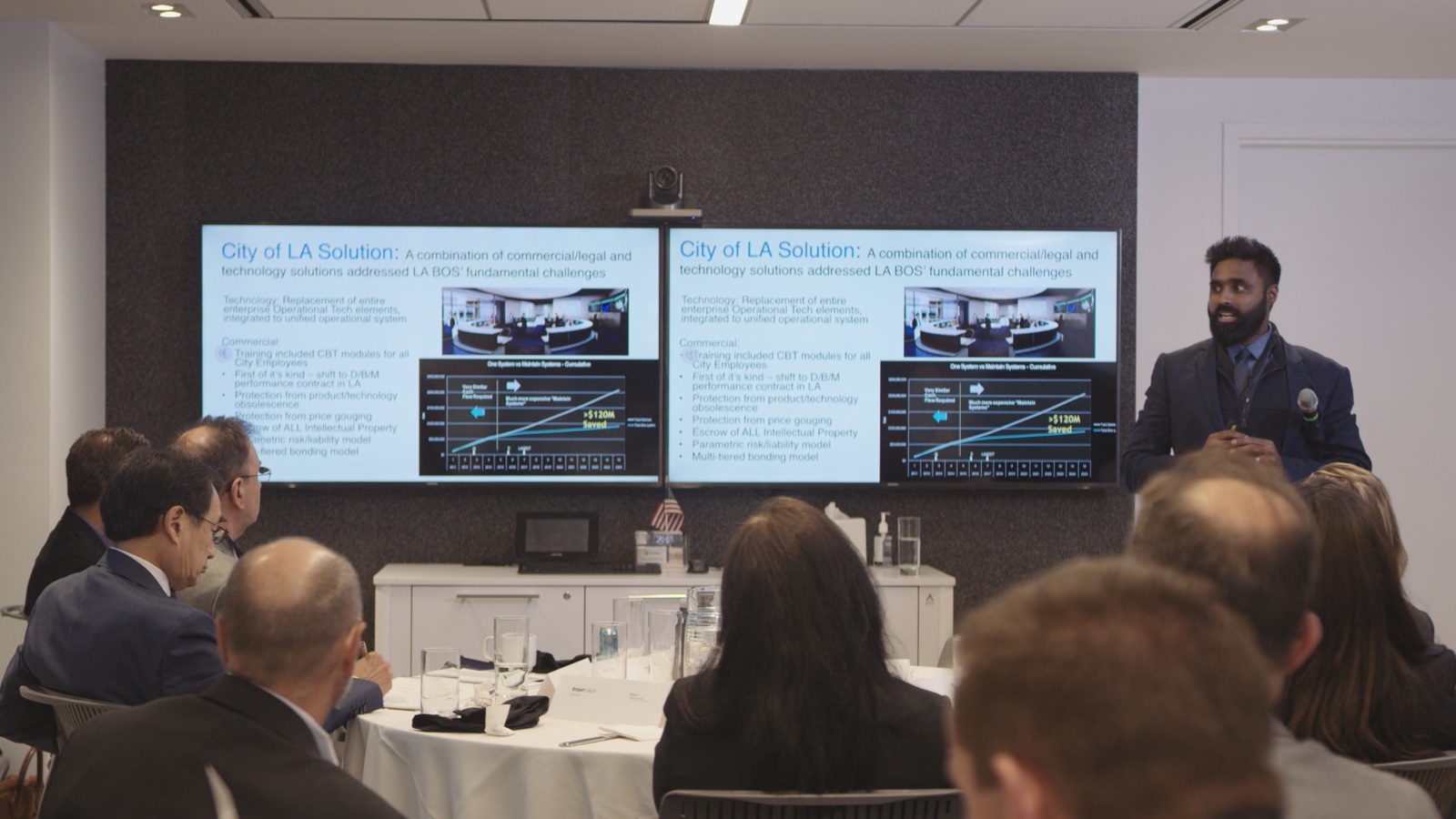 Livestream Man with short black hair and beard talking to a group of people in a video conference room