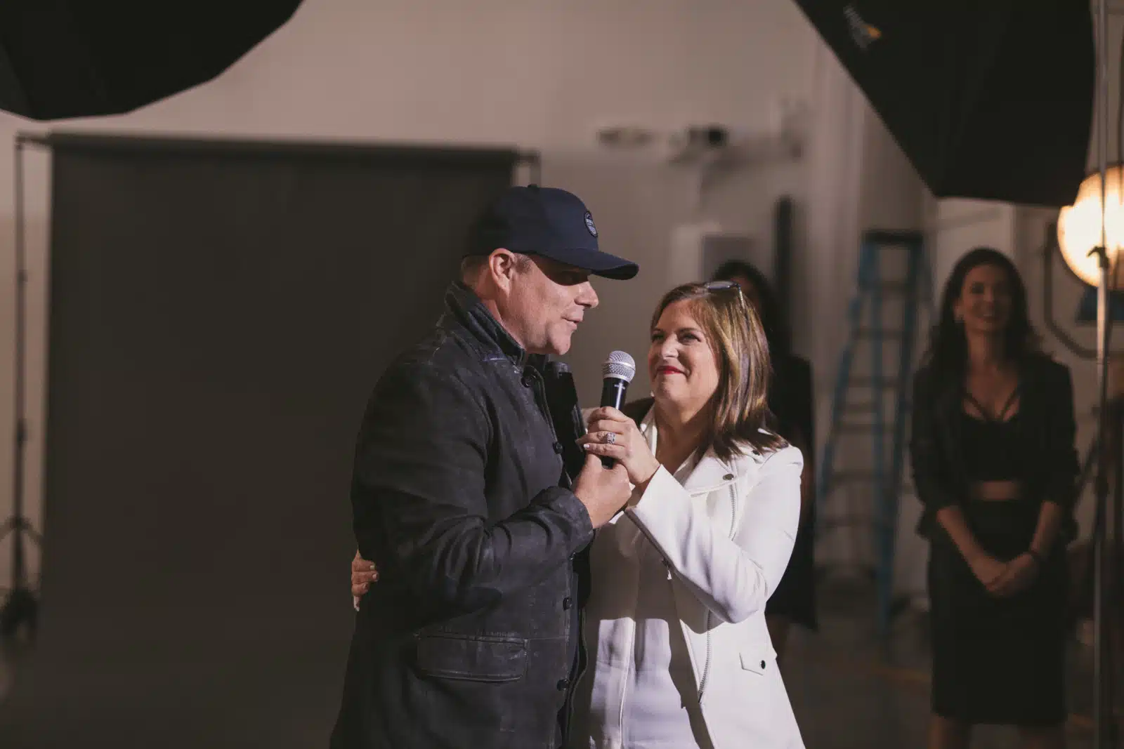 Man wearing black ball cap and jacket talking in microphone held by woman wearing white dress suit on set with two other women looking on in the background