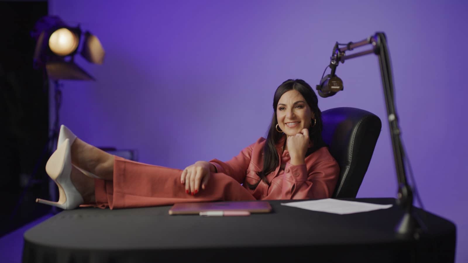 Woman with long black hair wearing an orange colored outfit and white heels posing for the camera smiling with her feet up on the table with a laptop, pen and piece of paper in front of her