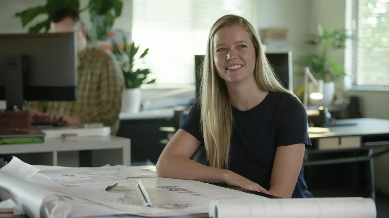 Headshot of woman with long blond hair sitting at a table with drafts smiling with a man working on a desktop in the background