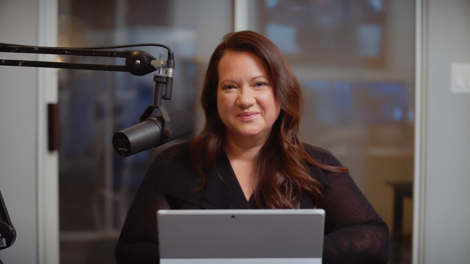 Keyword: Client Testimonial Videos Headshot of woman with long brown hair posing for the camera by a microphone smiling with a laptop in front of her.