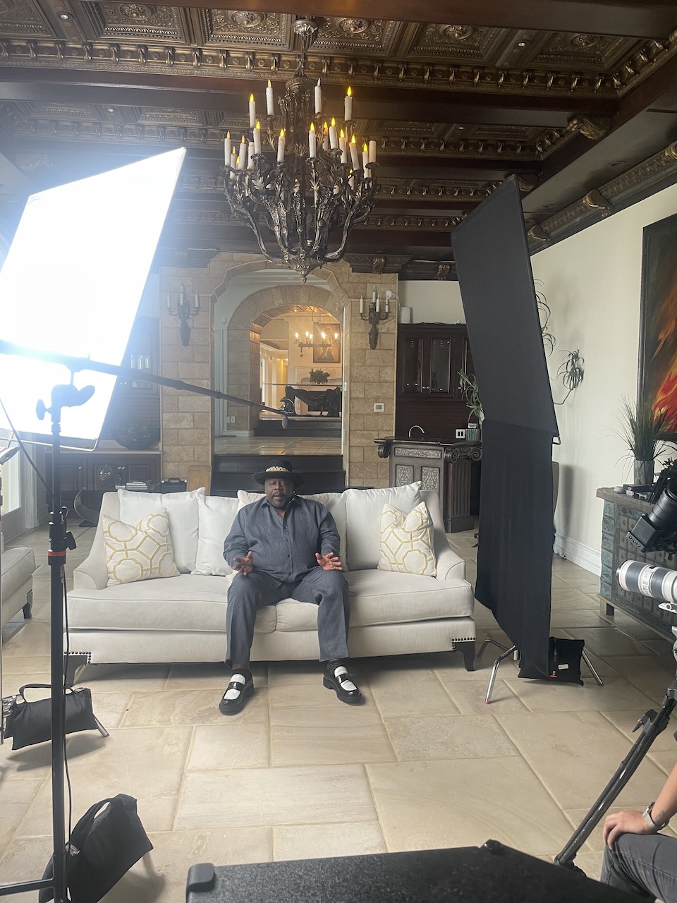 BlackPopBTS Cedric The Entertainer by C I Studios View of set with him sitting on the couch wearing a gray shirt and pants as well as a black hat talking to the camera