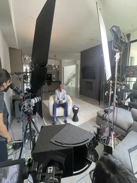 cis-page-video-production-partner-for-chinese-companies2-1 Steven A Smith wearing a light blue striped suit with dark blue pants and black shoes sitting on a white chair surrounded by equipment and crew