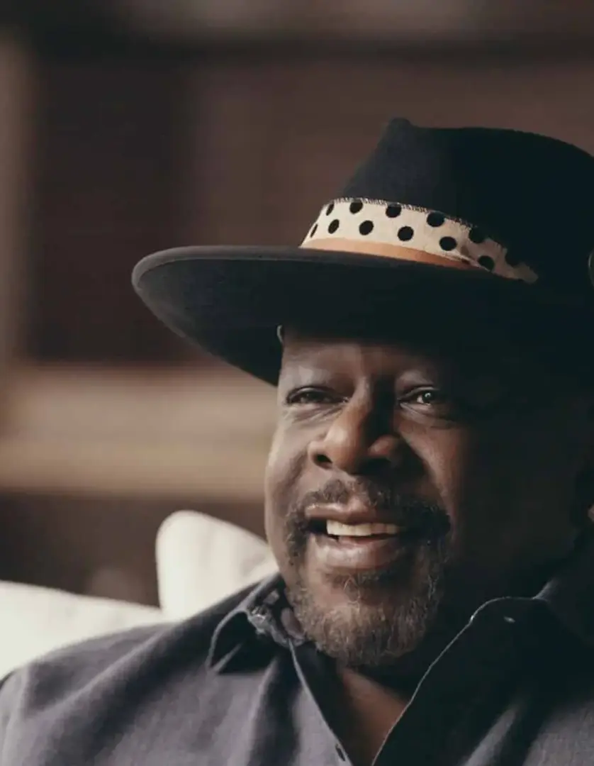 Black Pop 3 Cedric the Entertainer headshot sitting on a white couch smiling wearing a black hat and gray shirt
