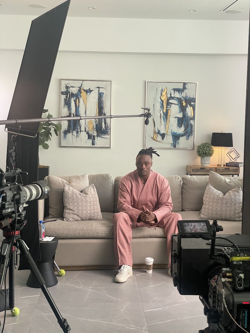 BlackPop Brandon Marsall sitting on a brown couch posing for the camera wearing a pink outfit with two video cameras trained on him