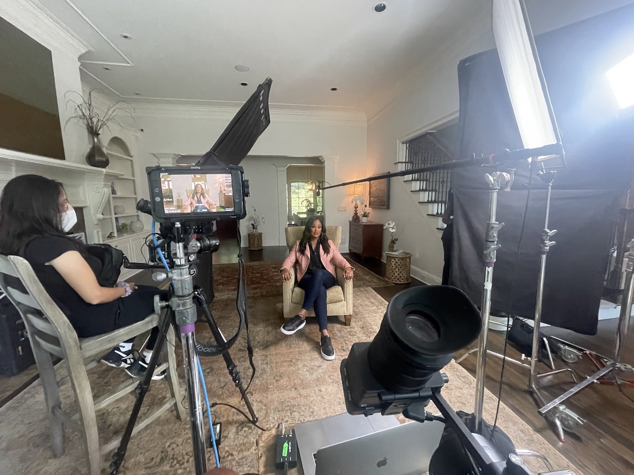 Leila Ali on set sitting on a beige chair wearing a pink dress jacket, black shirt, jeans and black sneakers with crew and equipment
