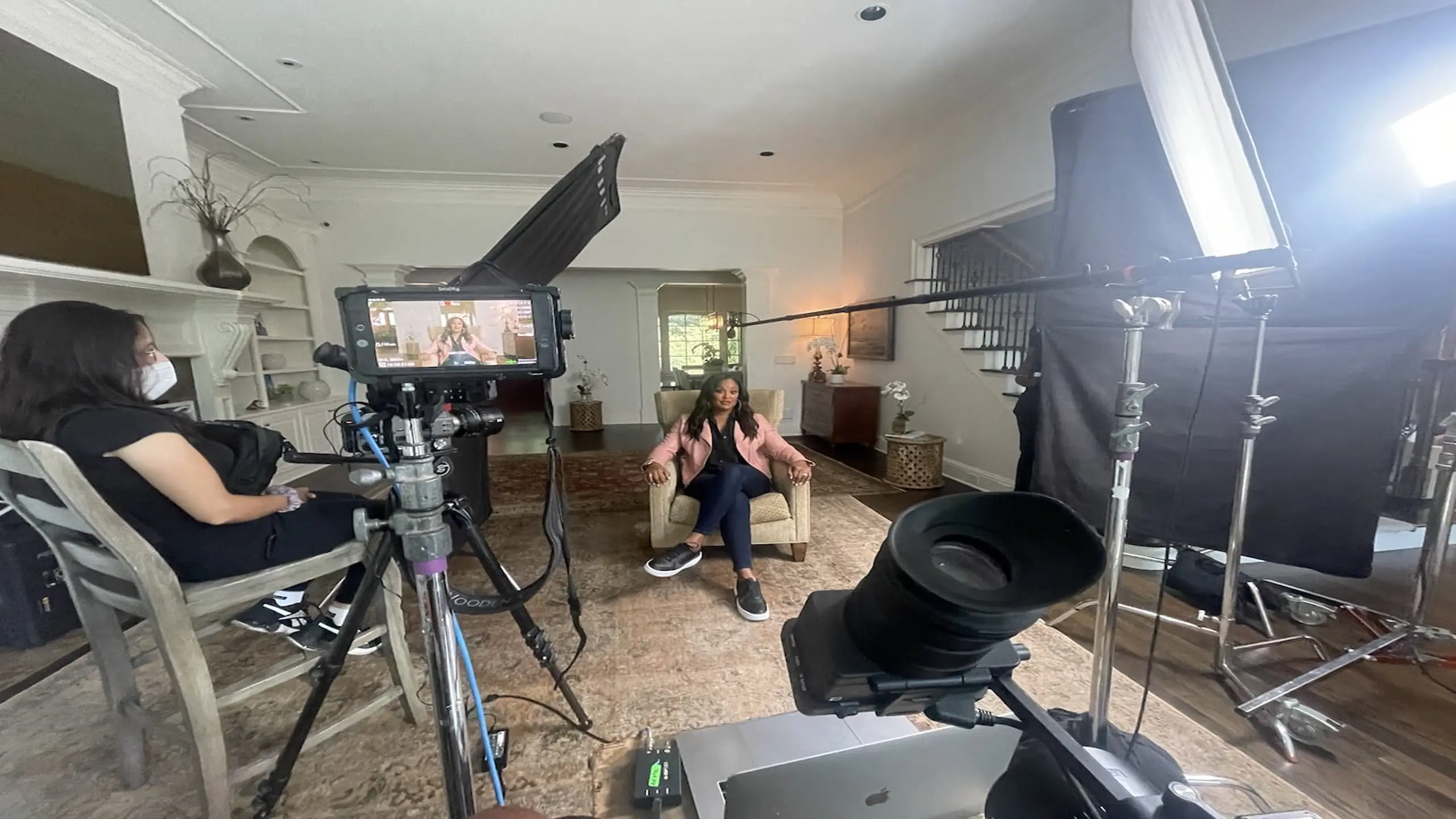 Leila Ali on set sitting on a beige chair wearing a pink dress jacket, black shirt, jeans and black sneakers with crew and equipment