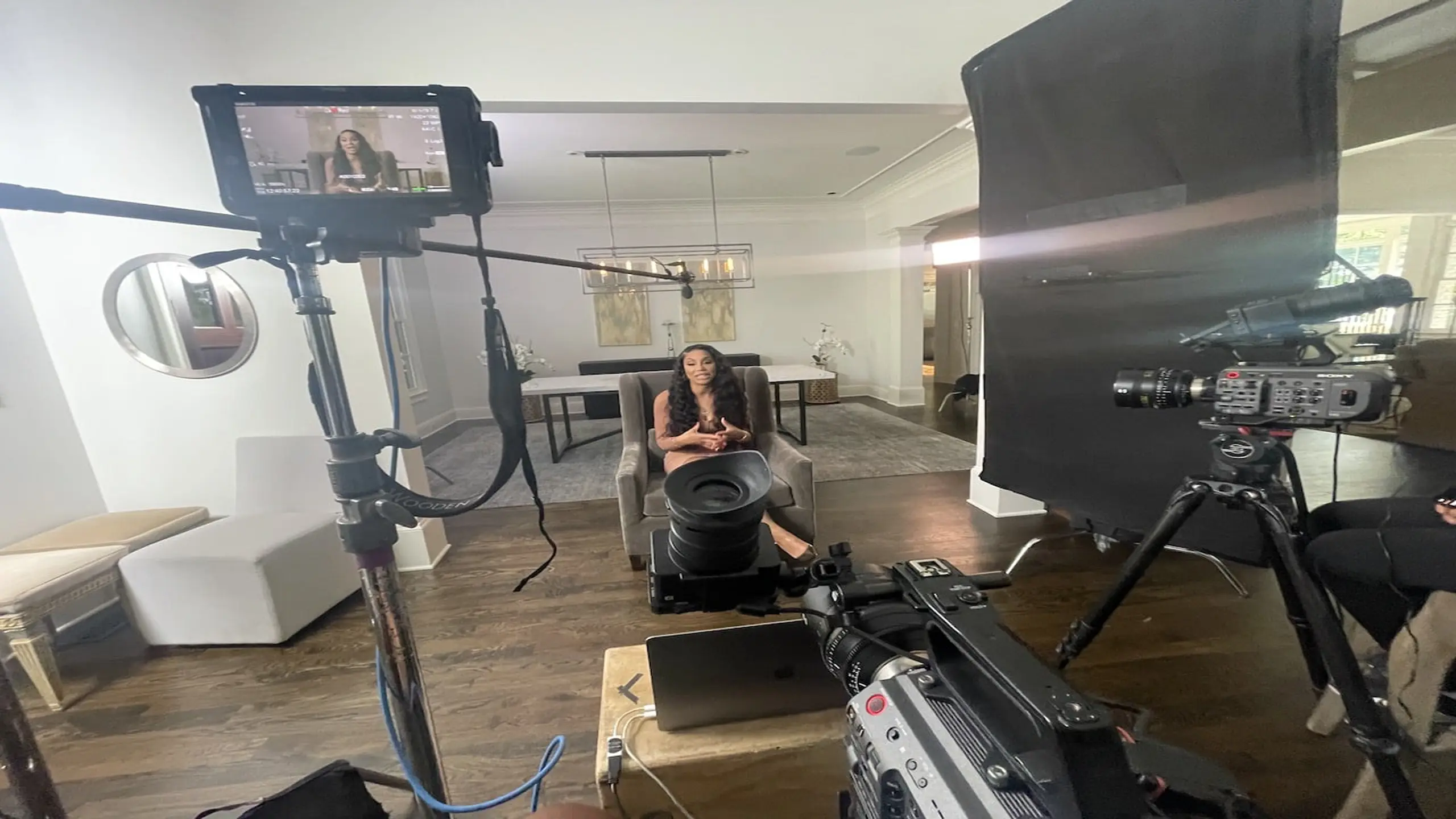 Tamar Braxton with long brown hair sitting on a gray chair talking with equipment around