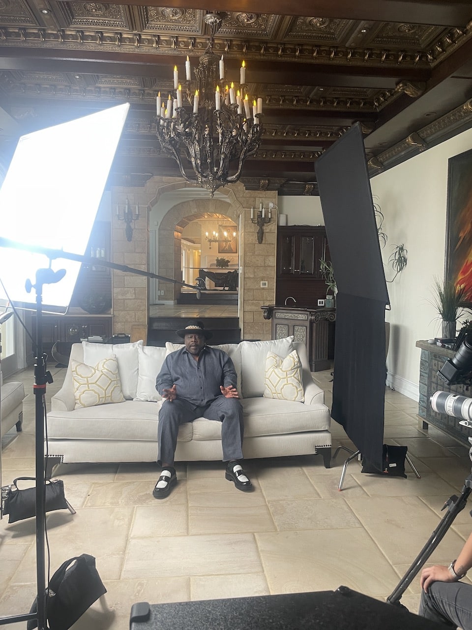 Cedric The Entertainer View of set with him sitting on the couch wearing a gray shirt and pants as well as a black hat talking to the camera