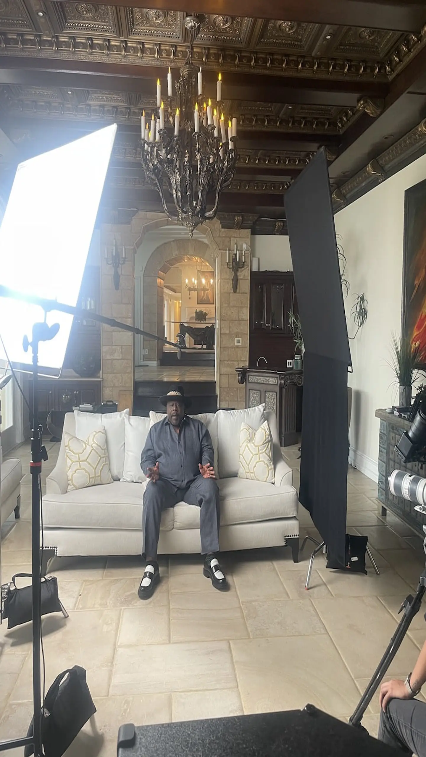 Cedric The Entertainer View of set with him sitting on the couch wearing a gray shirt and pants as well as a black hat talking to the camera