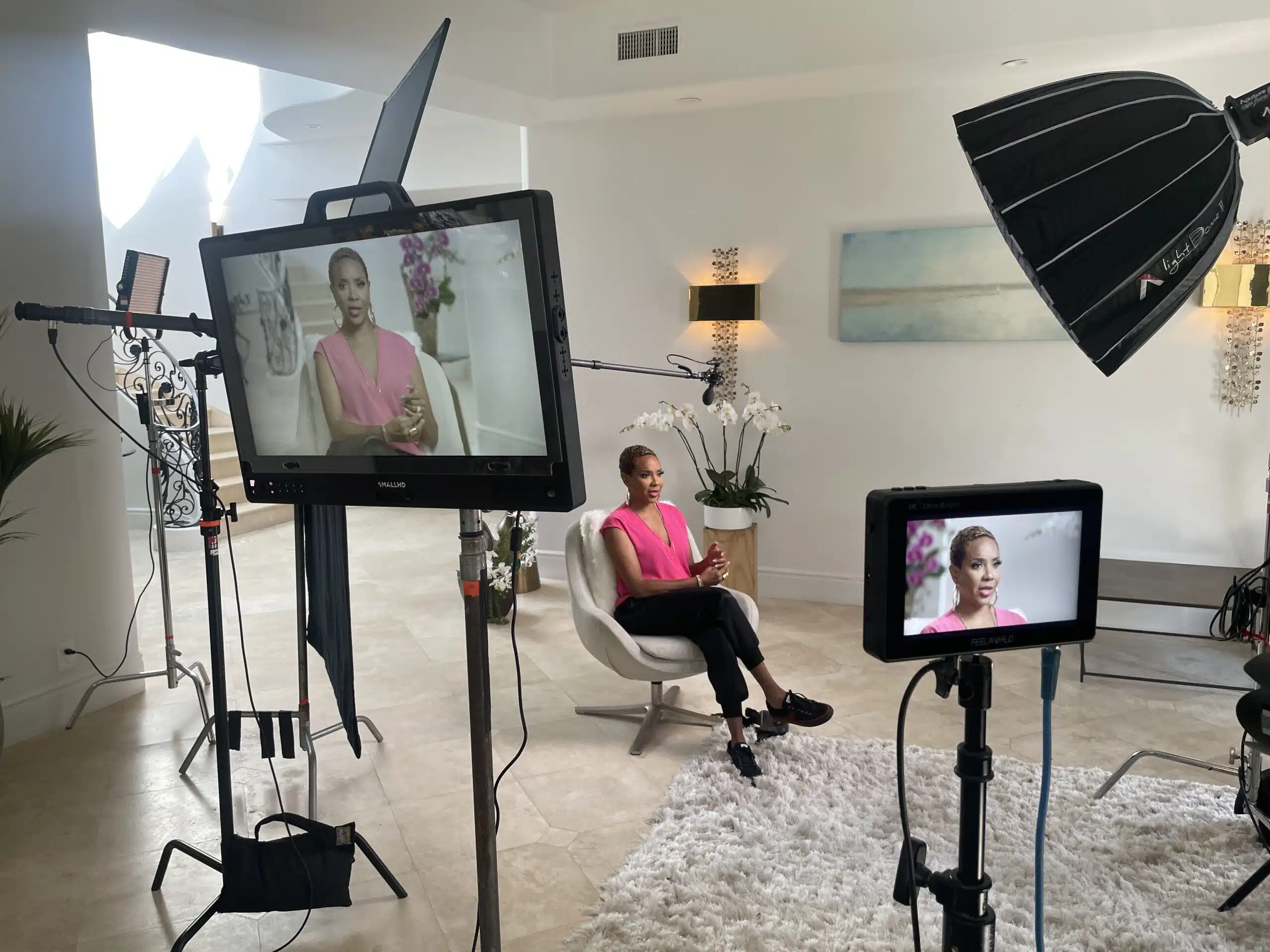 Closeup of video displays with MC Lyte sitting on a white chair wearing a pink dress top along with black pants and shoes