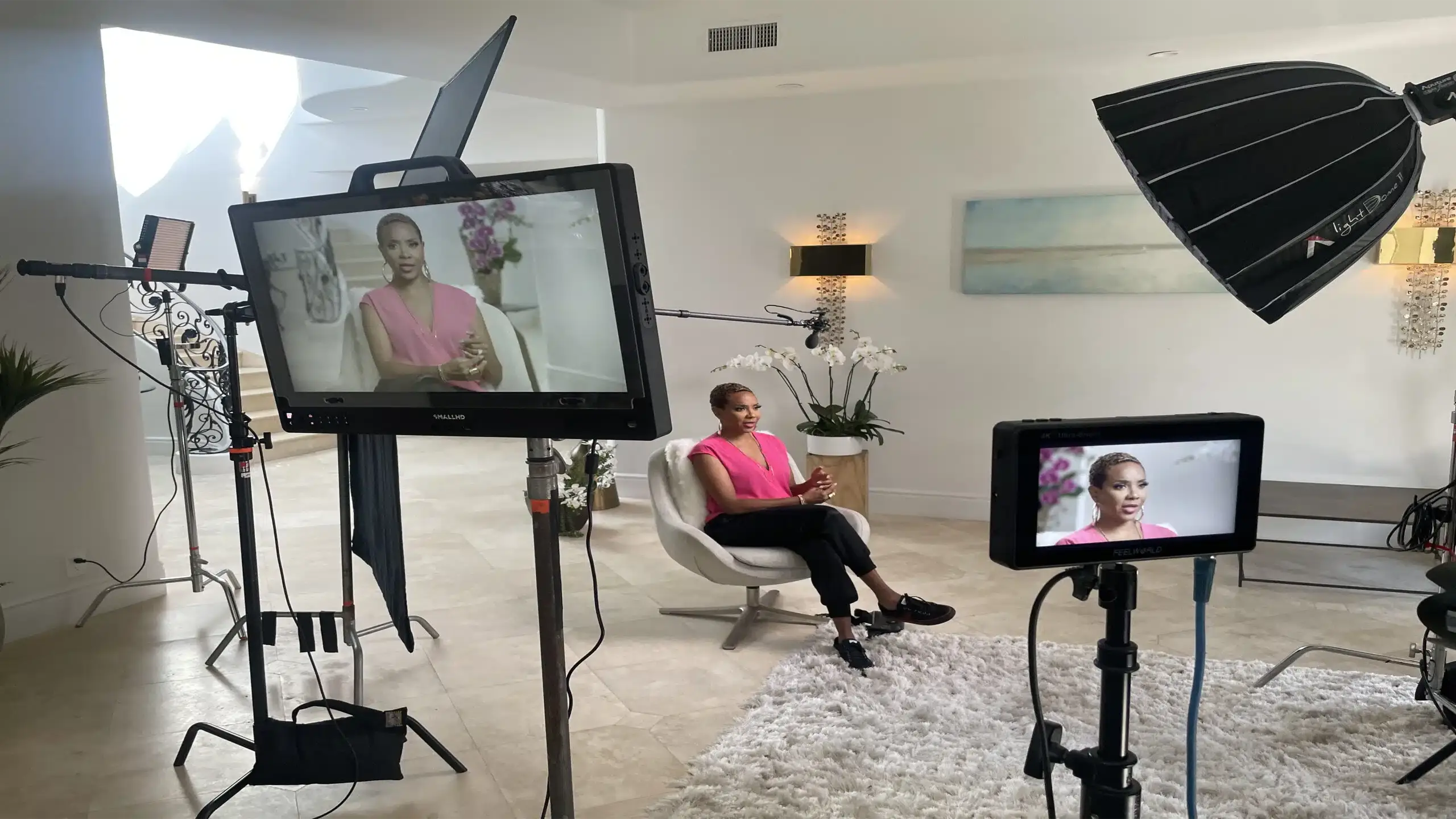Closeup of video displays with MC Lyte sitting on a white chair wearing a pink dress top along with black pants and shoes