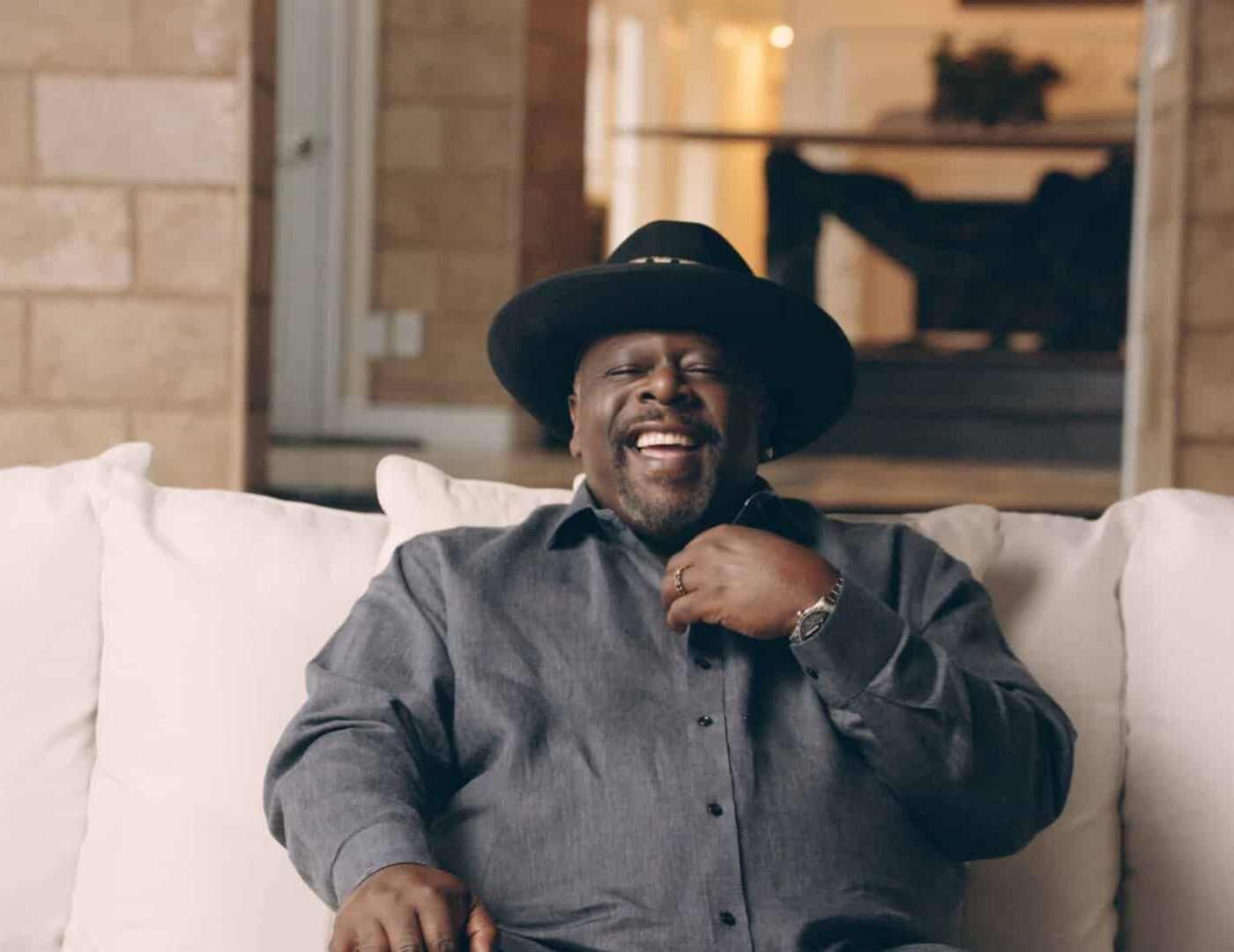 Cedric the Entertainer headshot sitting on a white couch smiling wearing a black hat and gray shirt