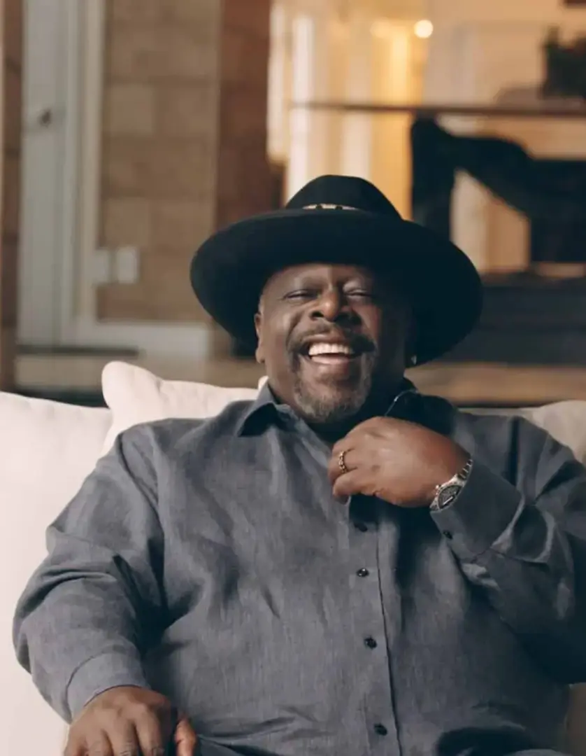 Black Pop 4 Cedric the Entertainer headshot sitting on a white couch smiling wearing a black hat and gray shirt