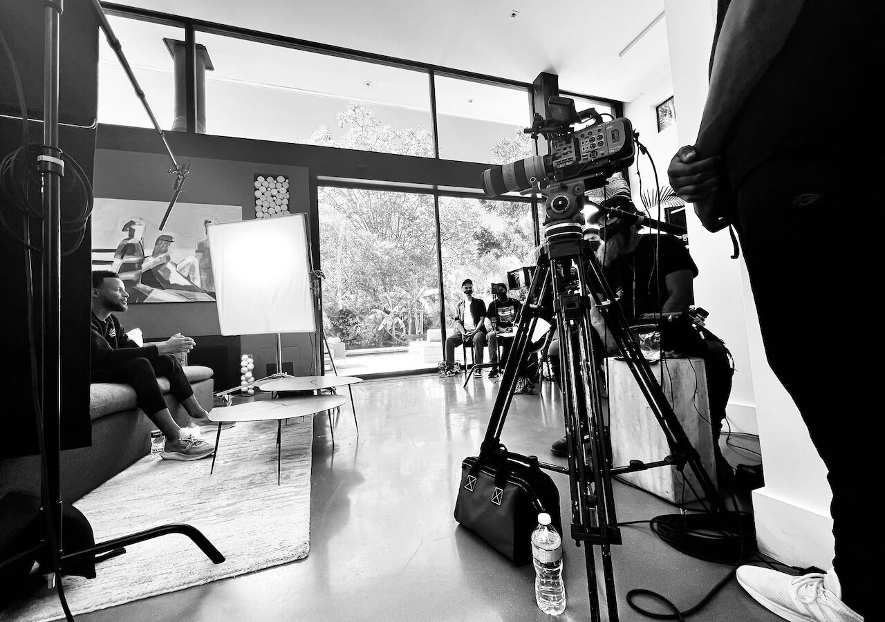 BlackPop Steph Curry with black and white of him on set sitting on a couch with tables near him along with crew and equipment