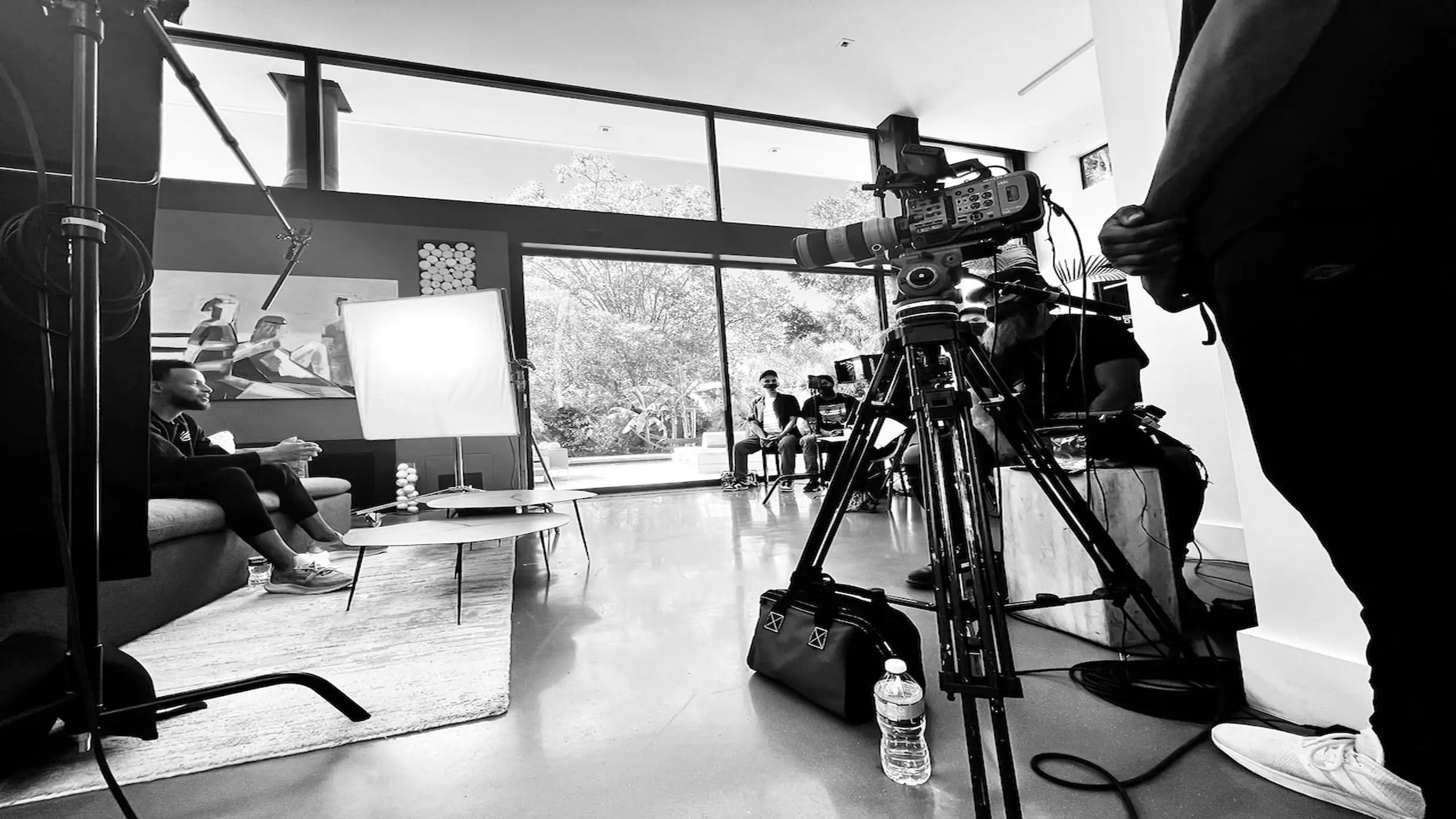 Black and white of Steph Curry on set sitting on a couch with tables near him along with crew and equipment