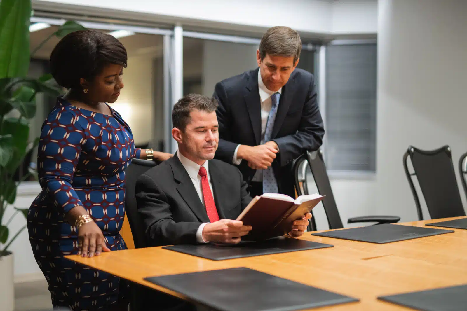 Man with a red tie sitting at a table holding a book with a man and woman behind him