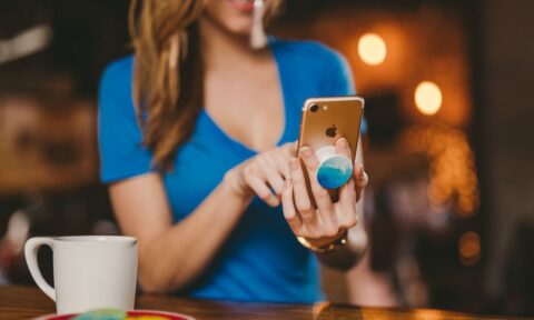 Woman wearing light blue shirt using a cell phone with white coffee cup and macaroons nearby