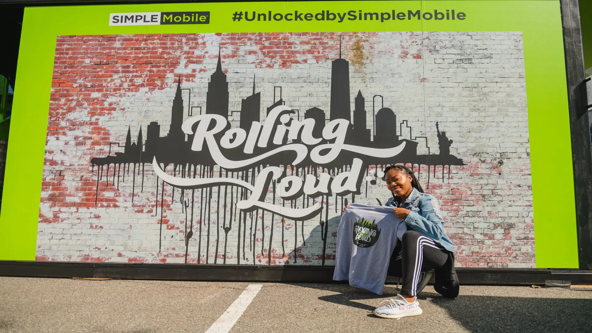 Rolling Loud 19 Woman holding up t shirt in front of Simple Mobile mural