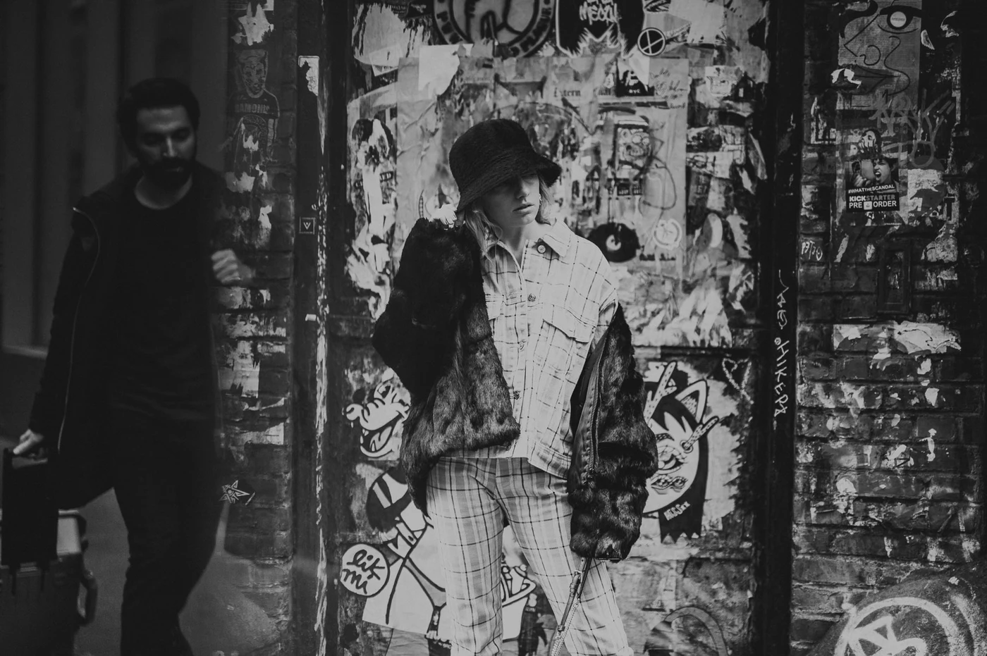 Black and white street photo of woman in fur coat and plaid against graffiti covered wall in New York City