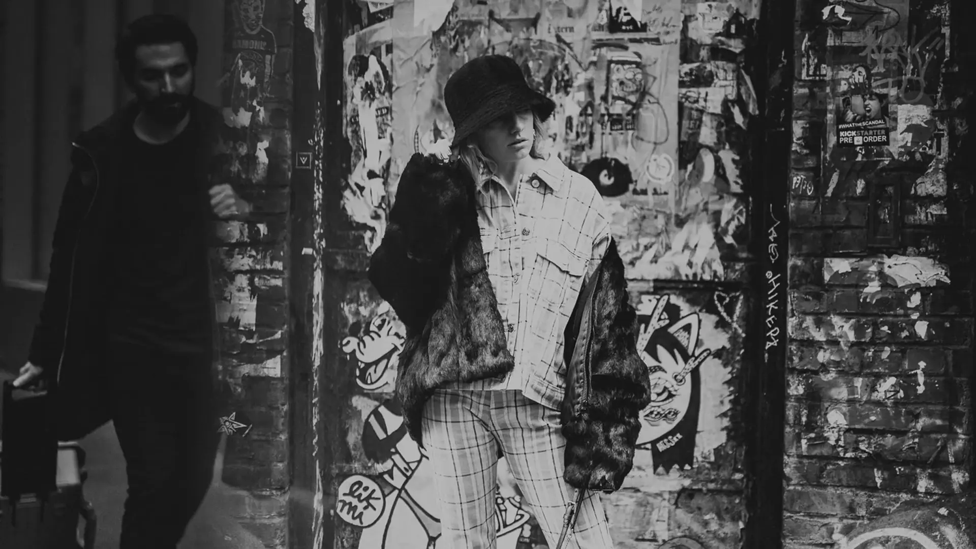 Black and white street photo of woman in fur coat and plaid against graffiti covered wall in New York City